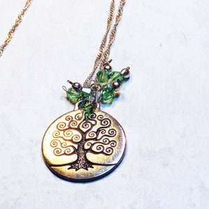May include: A gold-coloured necklace with a tree of life pendant. The pendant is circular with a detailed tree design. Green and silver beads are attached to the pendant. The necklace has a fine chain and a clasp.