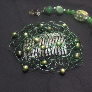 May include: A hair comb featuring a green wire mesh overlay adorned with small green beads, accompanied by a separate beaded strand with green stones and metal clips at each end. The design incorporates various shades of green throughout.