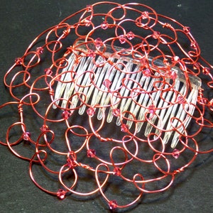 May include: A handcrafted wire kippah hair comb featuring a decorative design made of coral craft wire. The wire is intricately looped and adorned with small pink crystals. The comb itself is clear plastic, with evenly spaced teeth.