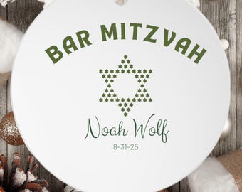 May include: A round white ceramic ornament featuring the green text BAR MITZVAH, a Star of David composed of small green stars, the name Noah Wolf in green script, and the date 8-31-25, suspended by a thin red ribbon.