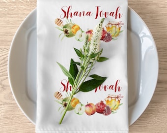 May include: White linen napkin with the text "Shana Tova" in red script, decorated with watercolor illustrations of apples, pomegranates, honey, and flowers. The napkin is placed on a white plate.
