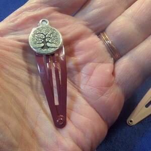 May include: Two hair barrettes. One is burgundy with a silver tree of life charm. The other is light pink with a silver charm. The barrettes are on a hand, with a blue background.