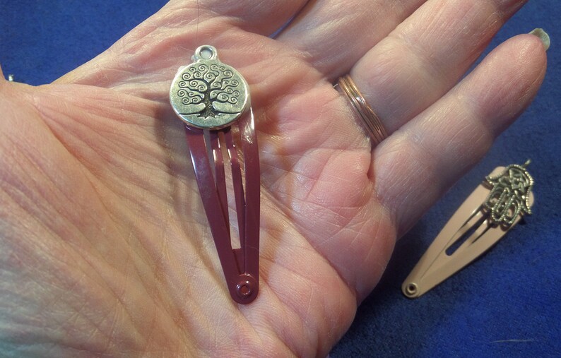 May include: Two hair barrettes. One is burgundy with a silver tree of life charm. The other is light pink with a silver charm. The barrettes are on a hand, with a blue background.