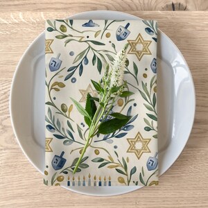 Puede incluir: Una servilleta de tela rectangular con un patr&oacute;n de acuarela de estrellas de David doradas, dreidels azules, ramas de olivo verdes y una fila de velas azules y doradas, rematada con un tallo de flor blanca sobre un plato blanco.