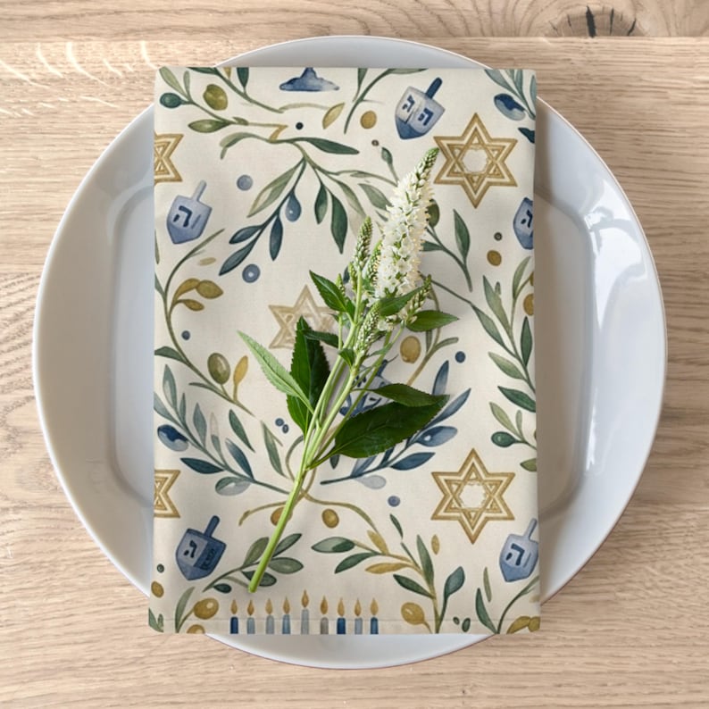 Puede incluir: Una servilleta de tela rectangular con un patr&oacute;n de acuarela de estrellas de David doradas, dreidels azules, ramas de olivo verdes y una fila de velas azules y doradas, rematada con un tallo de flor blanca sobre un plato blanco.