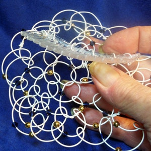 May include: A white wire mesh with gold and dark blue beads. The mesh is in the shape of a circle and is being held by a hand.