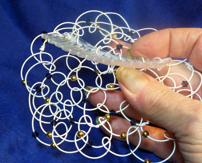 May include: A white wire mesh with gold and dark blue beads. The mesh is in the shape of a circle and is being held by a hand.