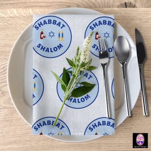 May include: A blue and white patterned cloth napkin with the words "Shabbat Shalom" repeated in a circle. The napkin is folded and placed on a white plate with two silver forks and a silver spoon.