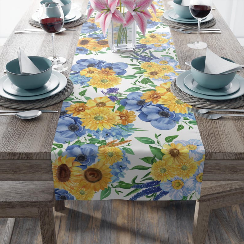 Image is of a Coastal Blue and Yellow Anemone and Daisy Floral tablecloth on a wood table background with plates and bowl table settings.
