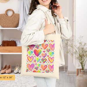 Peut inclure: Un sac fourre-tout en toile couleur cr&egrave;me mesurant 38 cm sur 41 cm, orn&eacute; d'un motif r&eacute;p&eacute;titif et vibrant de c&oelig;urs multicolores dans des tons de rose, jaune, bleu et orange, avec une esth&eacute;tique de style aquarelle sur le panneau avant.