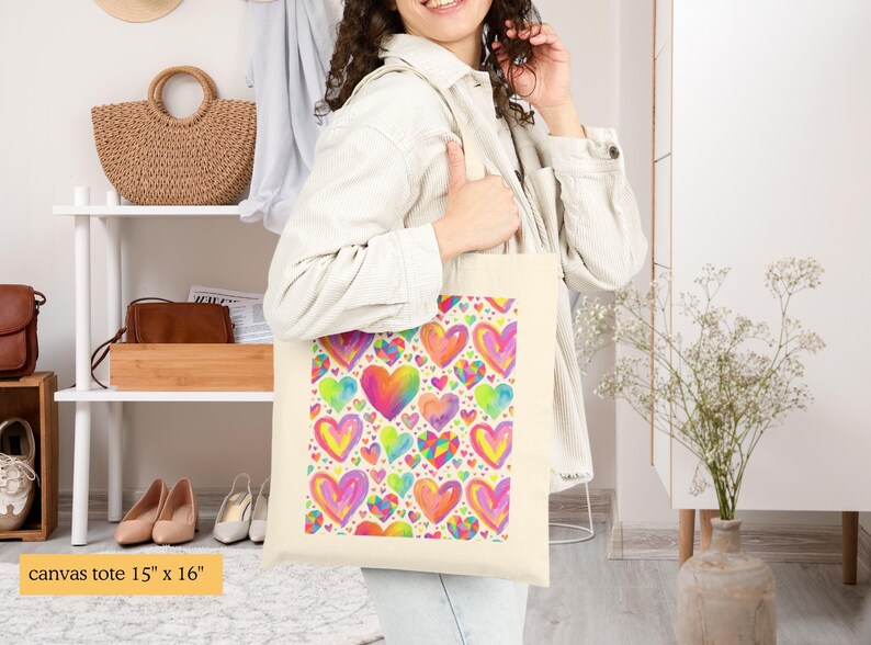 Peut inclure: Un sac fourre-tout en toile couleur cr&egrave;me mesurant 38 cm sur 41 cm, orn&eacute; d'un motif r&eacute;p&eacute;titif et vibrant de c&oelig;urs multicolores dans des tons de rose, jaune, bleu et orange, avec une esth&eacute;tique de style aquarelle sur le panneau avant.