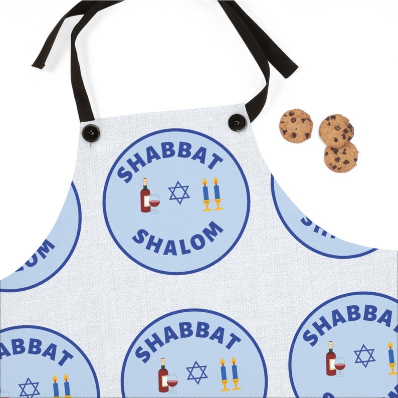 May include: A white apron with a blue circle pattern that says "Shabbat Shalom" with a wine glass, bottle of wine, and candles. The apron has black straps and two black buttons.