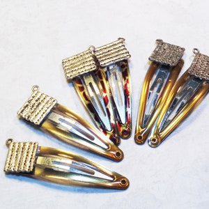 May include: Six hair barrettes with a gold-colored metal top and a tortoiseshell design. The barrettes are made of a clear, amber-colored material with a metal clip. The tops have a textured, rectangular design.