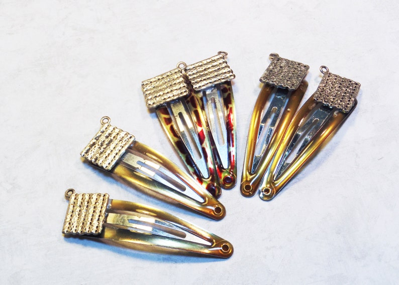 May include: Six hair barrettes with a gold-colored metal top and a tortoiseshell design. The barrettes are made of a clear, amber-colored material with a metal clip. The tops have a textured, rectangular design.