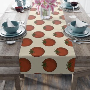 May include: A beige table runner featuring a repeating pattern of red tomatoes with green stems. The runner is placed on a wooden table set for a meal, with plates, glasses, and silverware visible.