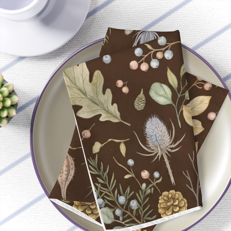 May include: A folded cloth napkin featuring a dark brown background with a repeating watercolor pattern of oak leaves, pinecones, thistles, small berries, and various botanical sprigs in muted shades of green, blue, and tan.