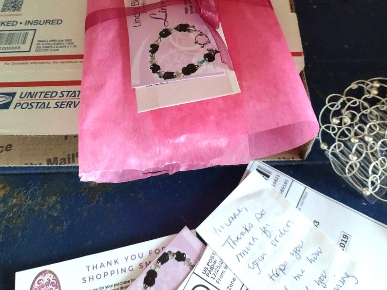 May include: A parcel wrapped in pink tissue paper, tied with a dark pink ribbon. A tag featuring a bracelet design is attached. A USPS box and a handwritten note are also visible.