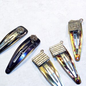 May include: A collection of six vintage hair barrettes. Two are black with star-shaped embellishments, and four have a colorful, patterned design with silver-toned metal accents. The barrettes are arranged on a light-colored surface.