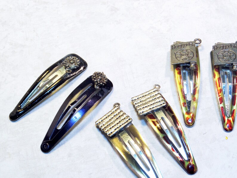 May include: A collection of six vintage hair barrettes. Two are black with star-shaped embellishments, and four have a colorful, patterned design with silver-toned metal accents. The barrettes are arranged on a light-colored surface.