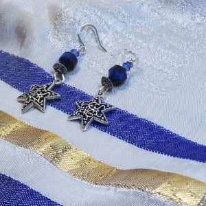 May include: A pair of silver-toned star earrings with intricate cut-out designs. Each earring features a dark blue bead and a silver hook. The earrings rest on a white and blue striped fabric with a gold ribbon.