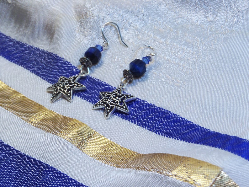 May include: A pair of silver-toned star earrings with intricate cut-out designs. Each earring features a dark blue bead and a silver hook. The earrings rest on a white and blue striped fabric with a gold ribbon.