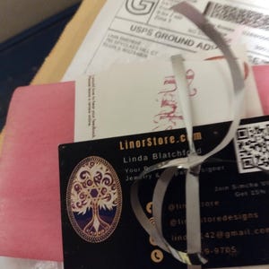 May include: A business card for LinorStore.com, a jewelry and pattern design business, featuring a tree of life design. The card is tied with a silver ribbon and is part of a package with a USPS shipping label.
