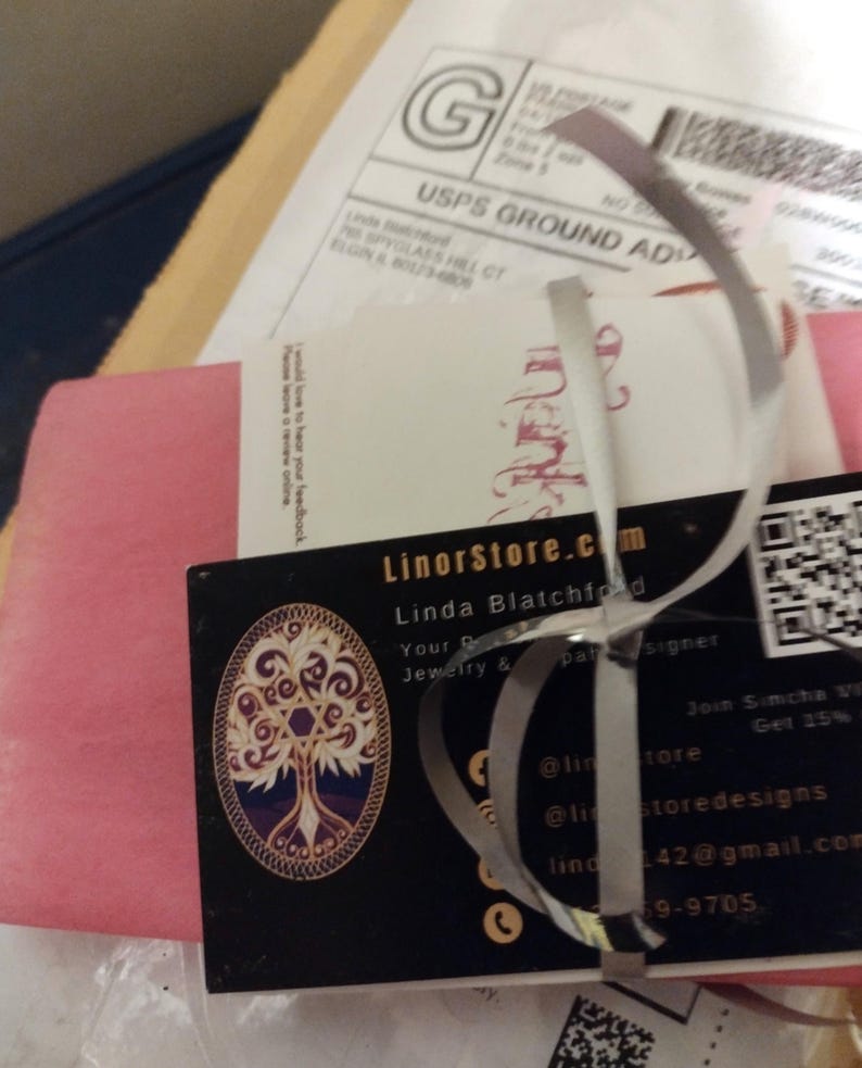 May include: A business card for LinorStore.com, a jewelry and pattern design business, featuring a tree of life design. The card is tied with a silver ribbon and is part of a package with a USPS shipping label.