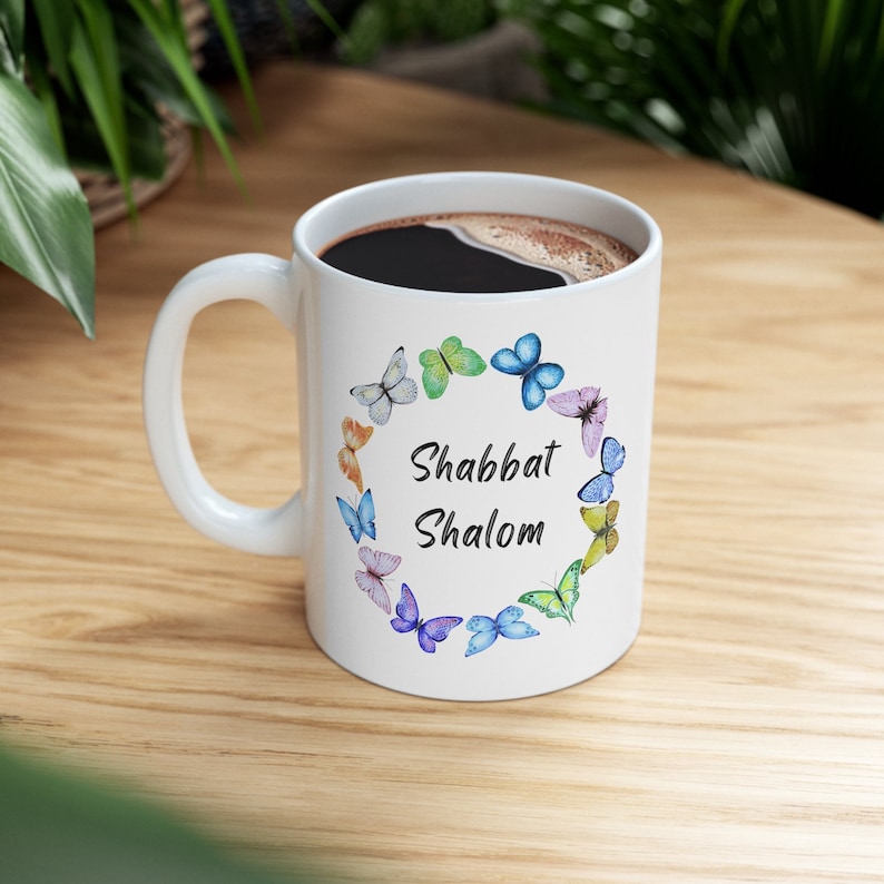 May include: White ceramic mug filled with dark liquid, featuring a colorful butterfly wreath design. The words "Shabbat Shalom" are centered within the wreath. The mug has a white handle and is set on a wooden surface.