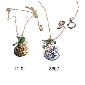 May include: Two necklaces featuring tree of life pendants. One pendant is gold-toned, the other silver-toned. Both have green bead accents and are on delicate silver chains. The necklaces are labelled T202 and 3807.