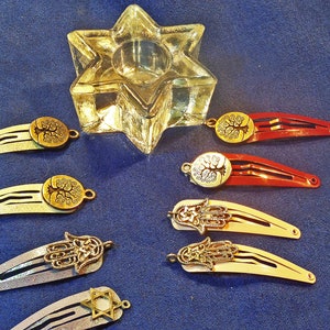 May include: A collection of hair barrettes with decorative charms, including a Star of David, a Hamsa hand, and a tree of life. The barrettes are in various colors, including red, gold, and silver. A clear glass star-shaped candle holder is also present.