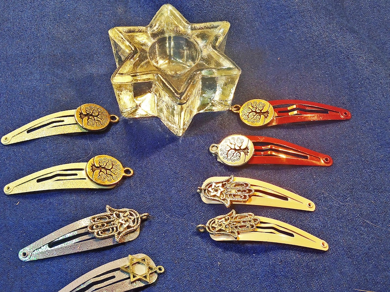 May include: A collection of hair barrettes with decorative charms, including a Star of David, a Hamsa hand, and a tree of life. The barrettes are in various colors, including red, gold, and silver. A clear glass star-shaped candle holder is also present.