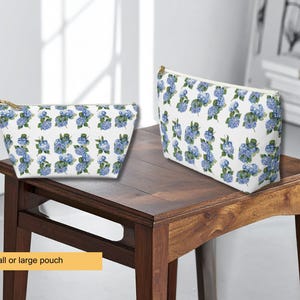 Puede incluir: Dos estuches con cremallera en tamaños pequeño y grande, con fondo blanco y un patrón repetitivo de racimos de flores de hortensia azules. Cada estuche tiene un cierre de cremallera dorado en el borde superior y una forma rectangular.