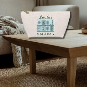 May include: A cream-colored zippered pouch featuring the text Linda's MAHJ BAG in dark green lettering. Below the name, a graphic displays eight light blue rectangular tiles with dark green line art illustrations of various mahjong symbols.