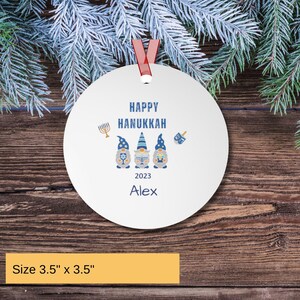 May include: A round white ornament measuring 3.5 inches by 3.5 inches featuring the text Happy Hanukkah, the year 2023, and the name Alex. The design includes three gnomes in blue and grey hats, a gold menorah, and a blue dreidel.