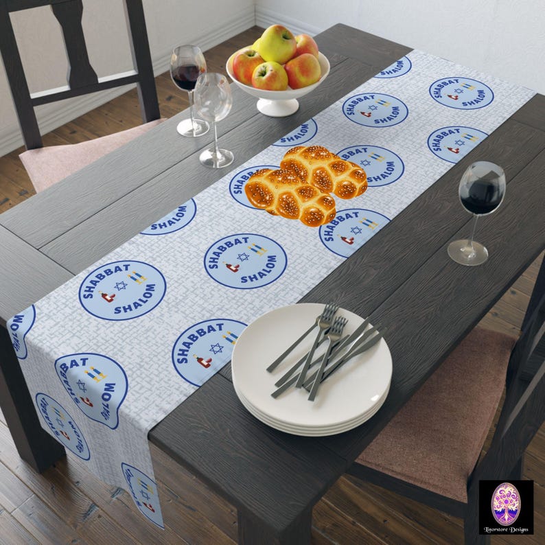 May include: A white table runner with a blue and white pattern featuring the Hebrew words "Shabbat Shalom" repeated throughout. The runner is laid on a dark wood table with a bowl of apples, two wine glasses, and a plate with silverware.
