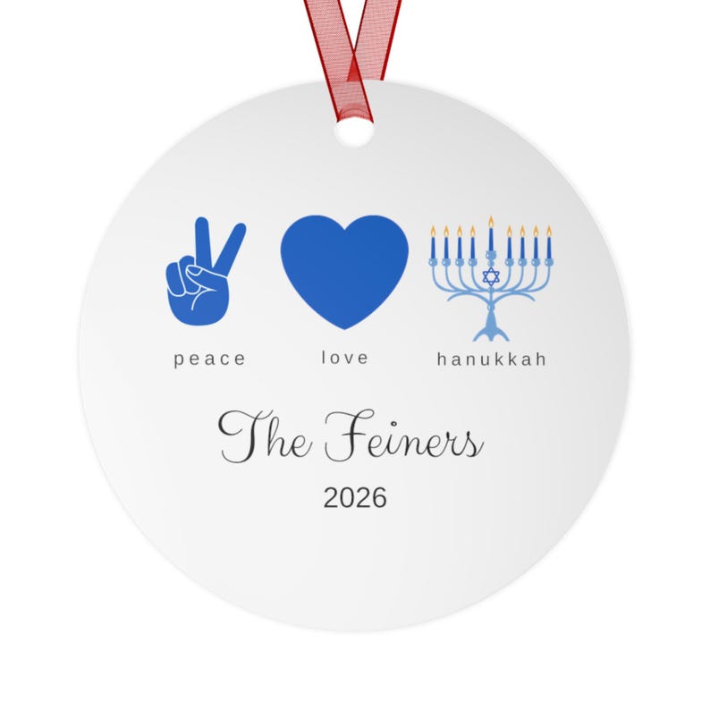 May include: A round white ornament with a red ribbon hanger featuring blue icons of a peace sign, a heart, and a menorah with the text peace, love, hanukkah, The Feiners, and 2026 printed in black and blue ink on the front surface.
