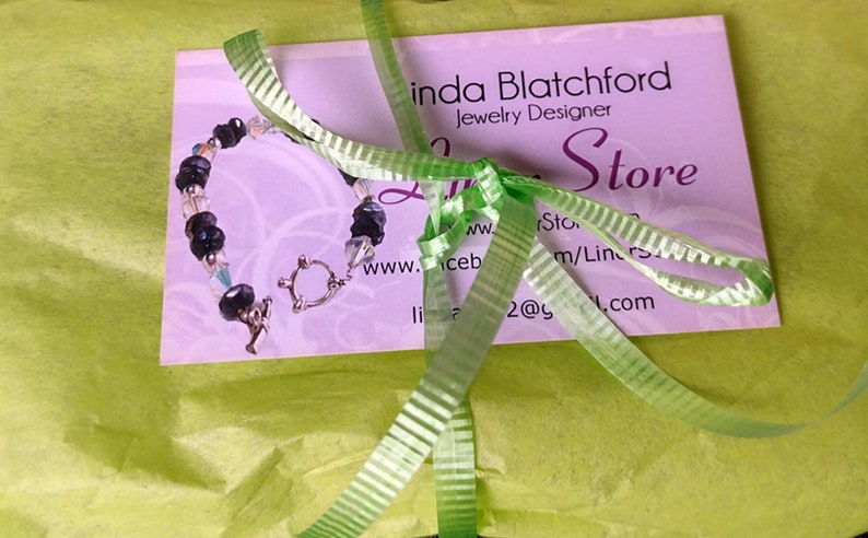 May include: A beaded bracelet with black and clear beads, fastened with a silver clasp, lies on a pink business card. The card is tied with a green ribbon and is on green tissue paper. The card reads "Linda Blatchford Jewelry Designer".