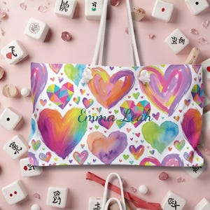 Peut inclure: Un sac fourre-tout blanc avec deux anses en corde blanche, orn&eacute; d'un motif color&eacute; de c&oelig;urs peints et g&eacute;om&eacute;triques dans des tons de rose, violet, orange, jaune et vert. Le pr&eacute;nom Emma Leah est imprim&eacute; en &eacute;criture cursive bleu fonc&eacute; au centre.