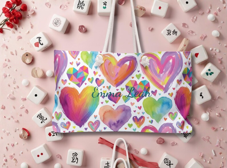 Peut inclure: Un sac fourre-tout blanc avec deux anses en corde blanche, orn&eacute; d'un motif color&eacute; de c&oelig;urs peints et g&eacute;om&eacute;triques dans des tons de rose, violet, orange, jaune et vert. Le pr&eacute;nom Emma Leah est imprim&eacute; en &eacute;criture cursive bleu fonc&eacute; au centre.