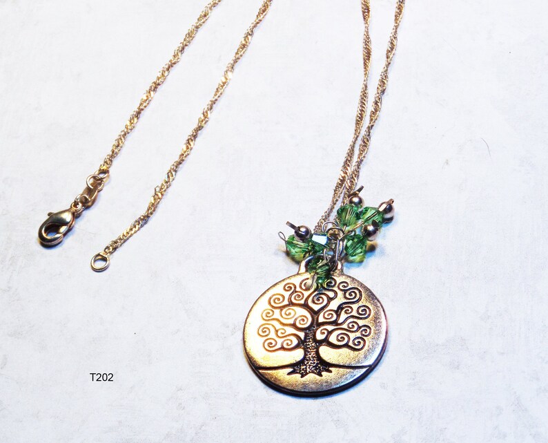 May include: A gold-coloured necklace with a tree of life pendant. The pendant is circular with a detailed tree design. Green and silver beads are attached to the pendant. The necklace has a fine chain and a clasp.