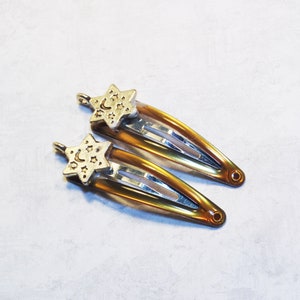 May include: Two hair barrettes with a gold star and moon charm. The barrettes are a warm amber color with silver metal clasps. The star charms have a crescent moon and star design.