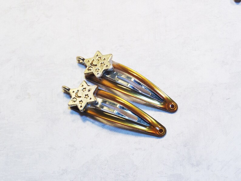 May include: Two hair barrettes with a gold star and moon charm. The barrettes are a warm amber color with silver metal clasps. The star charms have a crescent moon and star design.