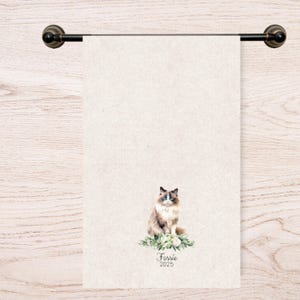Op de afbeelding: Een beige handdoek met een aquarel illustratie van een Ragdoll kat met blauwe ogen, zittend op een bed van groen. De handdoek hangt aan een donker bronzen handdoekrek. De naam "Fossie" en het jaar "2025" zijn eronder gedrukt.