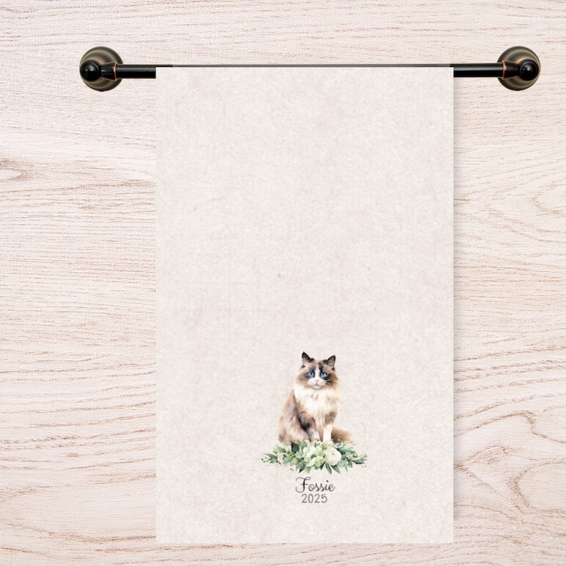 Op de afbeelding: Een beige handdoek met een aquarel illustratie van een Ragdoll kat met blauwe ogen, zittend op een bed van groen. De handdoek hangt aan een donker bronzen handdoekrek. De naam "Fossie" en het jaar "2025" zijn eronder gedrukt.