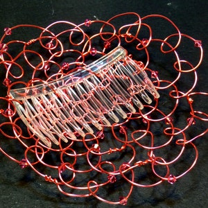 May include: A decorative hair comb featuring a clear plastic comb set within a frame of red wire. The wire is shaped into looping swirls and embellished with small pink beads. This handcrafted hair accessory adds a touch of elegance.