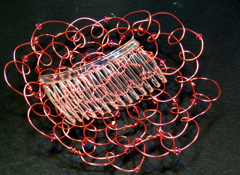 May include: A decorative hair comb featuring a clear plastic comb set within a frame of red wire. The wire is shaped into looping swirls and embellished with small pink beads. This handcrafted hair accessory adds a touch of elegance.