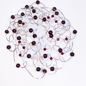 A silver-toned wire kippah with a tangled, openwork design. The necklace is adorned with dark red, round beads and smaller, faceted red crystals. The beads and crystals are interspersed throughout the wire loops, creating a visually interesting yarmulke.