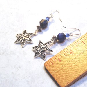 May include: A pair of silver-tone star earrings with intricate filigree detailing. Each earring features a star pendant, dark blue faceted beads, and small blue crystal accents. The earrings are displayed on a light-colored surface.