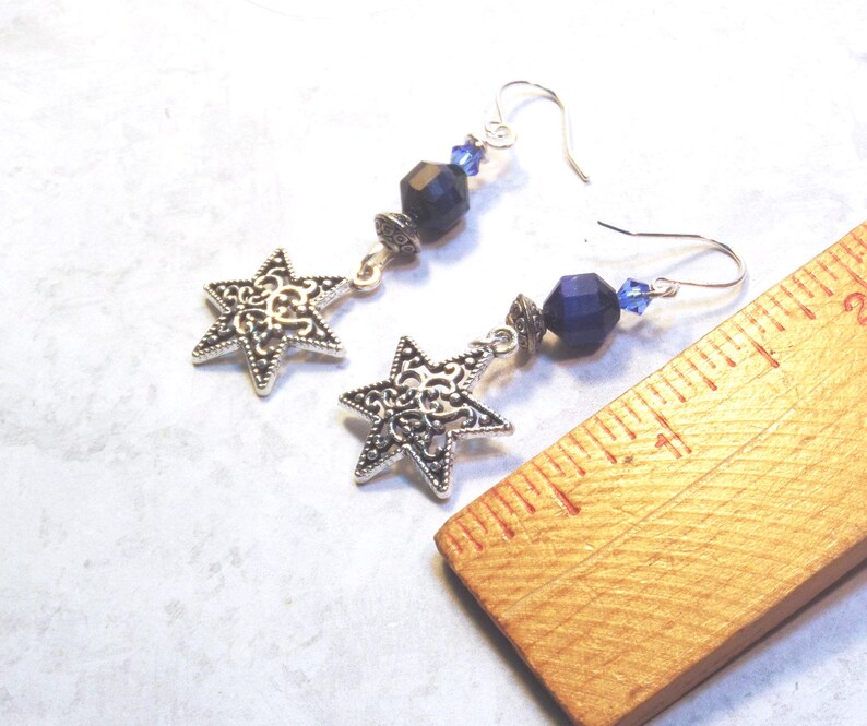 May include: A pair of silver-tone star earrings with intricate filigree detailing. Each earring features a star pendant, dark blue faceted beads, and small blue crystal accents. The earrings are displayed on a light-colored surface.