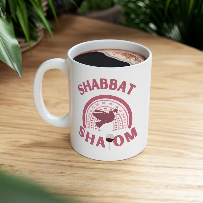 Image is of White ceramic coffee mug with a pink design featuring the text "Shabbat Shalom" and a dove with a star on its head flying over a rainbow. A glass of wine is the L in Shalom.
